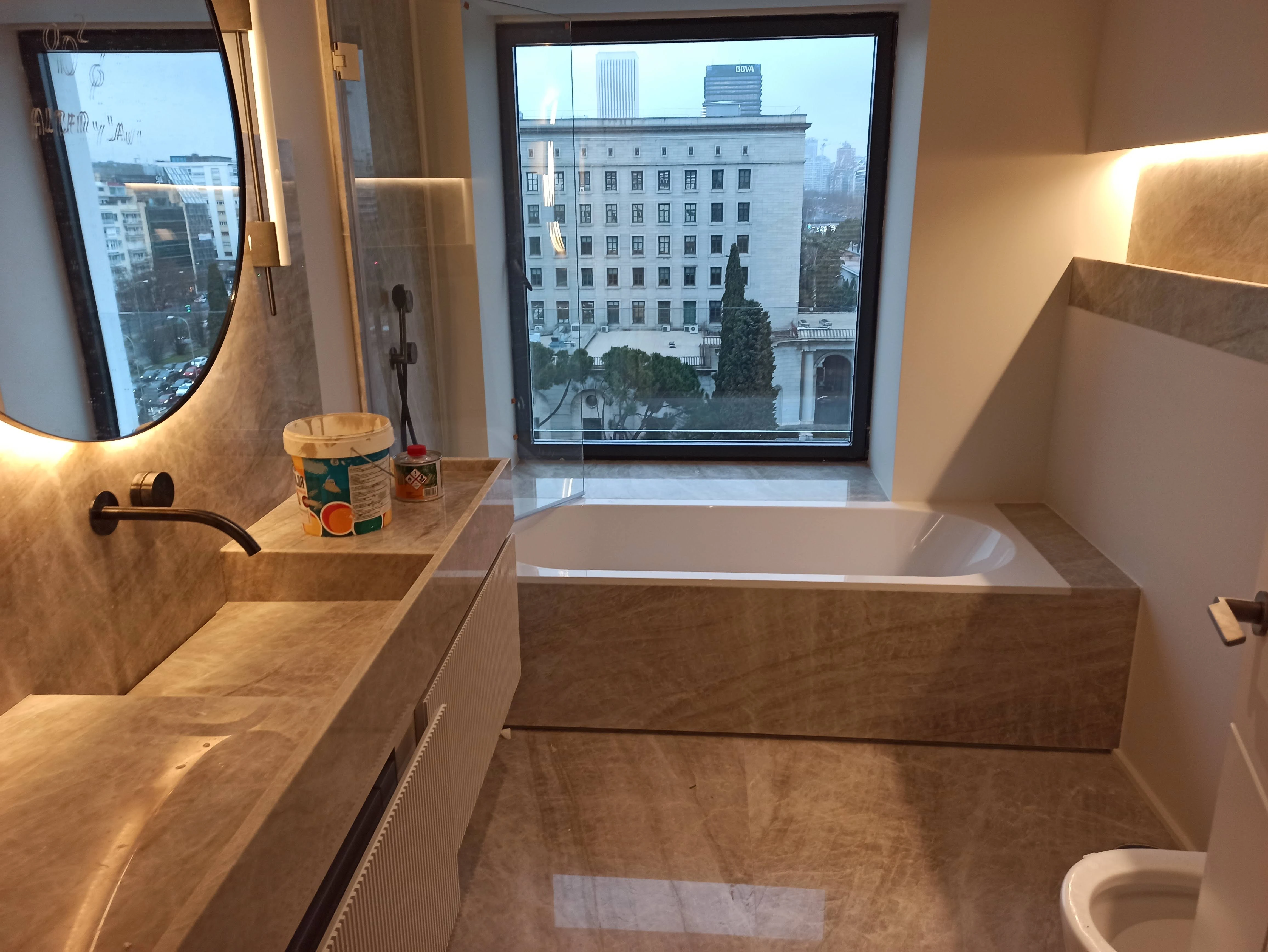 Baño urbano con bañera integrada en mármol beige y vistas a la ciudad - Ropema Rubio
